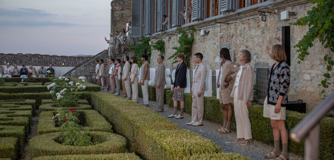 Männliche Models präsentieren Mode und stehen nebeneinander in einem Garten vor einem alten Haus