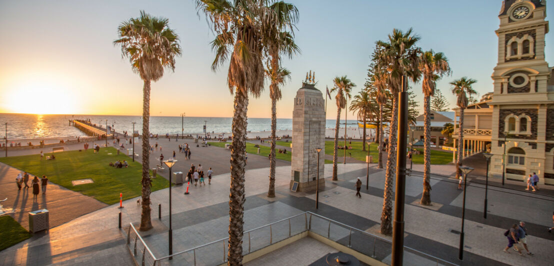 Ein Platz mit Palmen und einem Denkmal aus Stein vor dem Meer in Glenelg nahe Adelaide