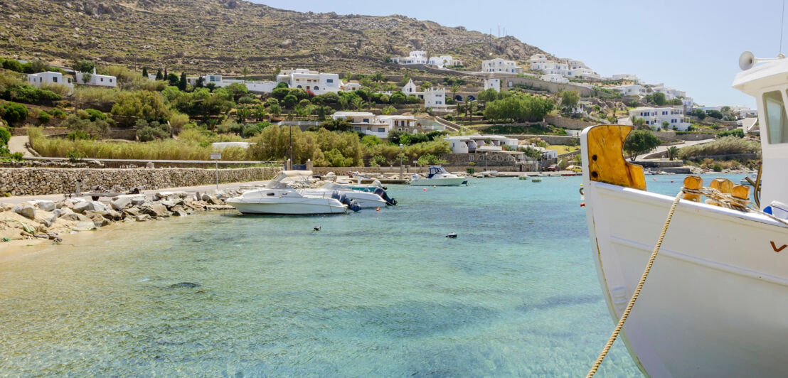 Fischerboote liegen an einem Strand von Mykonos, im Hintergrund weiß getünchte Häuser am Hügelhang