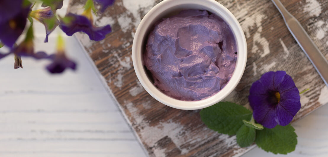 Schälchen mit lilafarbener Creme, angerichtet auf einem Holzbrett mit Blumen