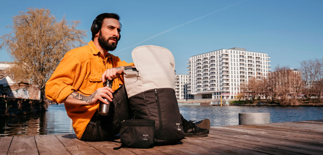 Ein am Wasser sitzender Mann mit geöffnetem Rucksack neben sich