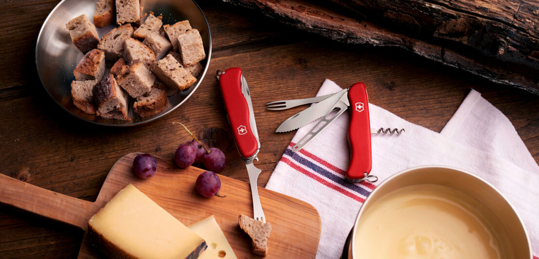 Zwei aufgeklappte, rote Schweizer Taschenmesser mit Käsemesser und Fonduegabel auf einem Holztisch neben Käsescheiben und einem Fonduetopf