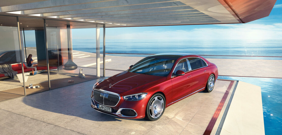 Ein roter Mercedes-Maybach seitlich von vorne am Pool auf der Terrasse eines modernen Hauses mit viel Glas, im Hintergrund blaues Meer