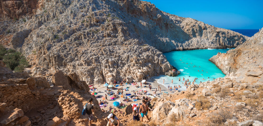 Blick auf einen zwischen Felsen liegenden Sandstrand, der einen schmalen Zugang zum türkisfarbenen Meer bietet