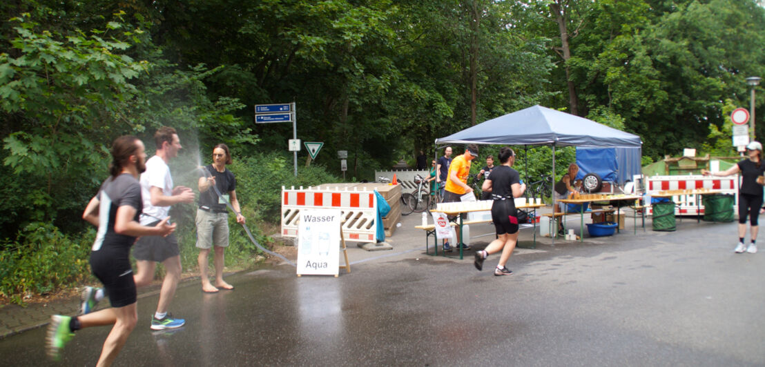 Mehrere Personen joggen an einem kleinen Stand vorbei und werden mit einem Wasserschlauch nassgespritzt