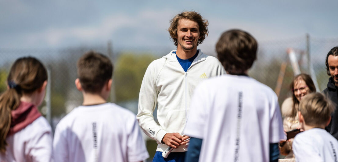 Alexander Zverev spricht zu Kindern auf einem Tennisplatz.