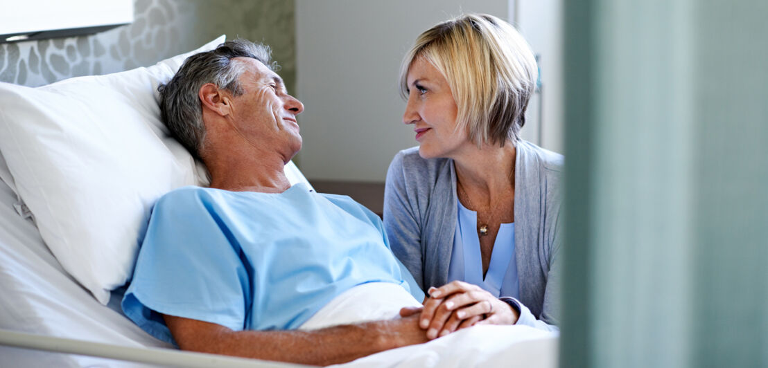 Ein Mann in einem Krankenbett, daneben eine Frau, die ihm die Hände hält