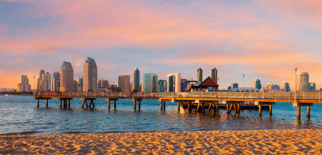 Skyline von San Diego mit einem Pier am Sandstrand im Vordergrund bei Sonnenuntergang