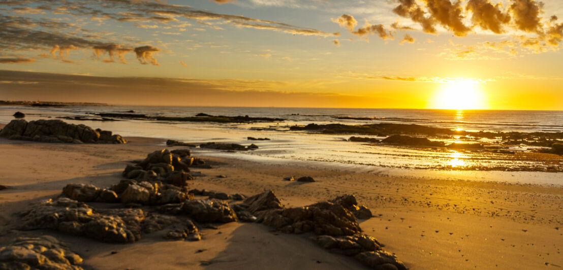 Ein menschenleerer, teilweise felsiger Sandstrand mit Sonnenuntergang über dem Meer