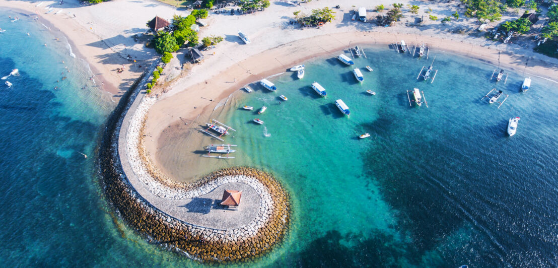 Luftaufnahme eines Küstenabschnittes mit Booten im türkisblauen Wasser vor einem weißen Sandstrand mit einem geschwungenen Wellenbrecher