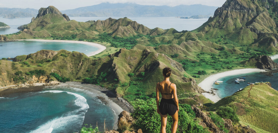 Eine Person steht auf einem Felsvorsprung und blickt über einen Teil des Komodo-Nationalparks mit kleinen Bergen und Buchten