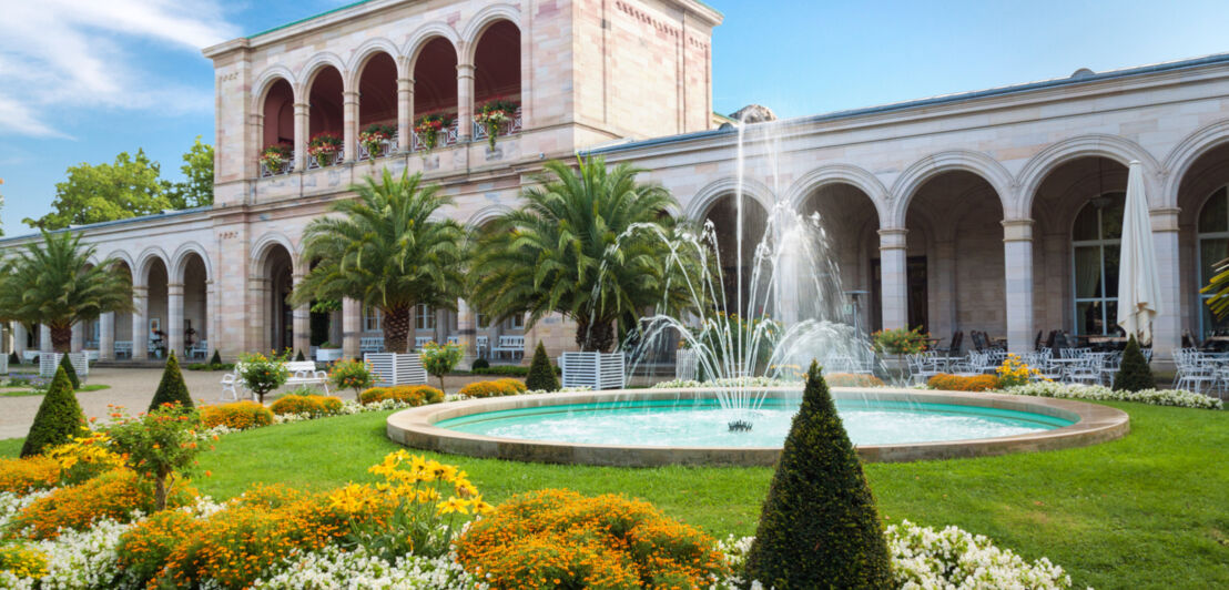 Wandelhalle mit Säulen in Bad Kissingen, im Vordergrund ein Park mit Palmen und Springbrunnen