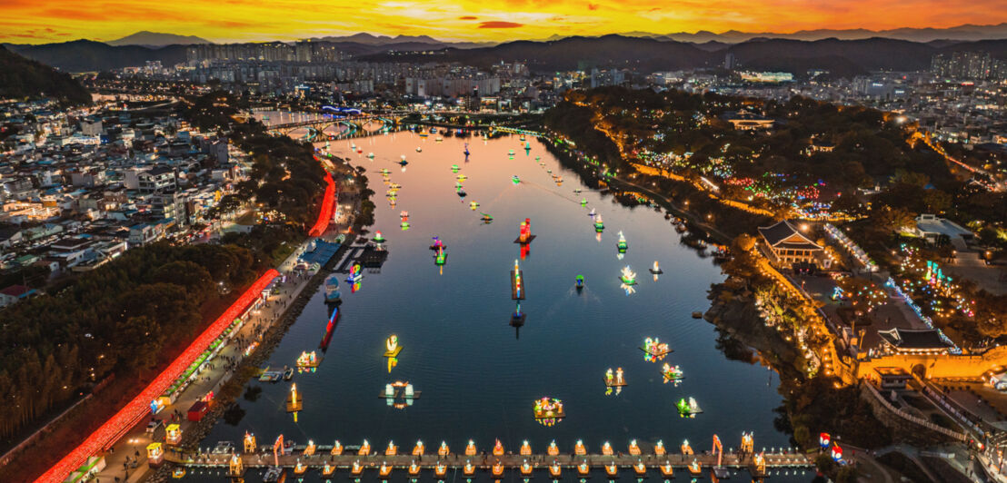 Luftaufnahme der Stadt Jinju mit einem breitem Fluss, auf dem bunte Laternen schwimmen bei Sonnenuntergang