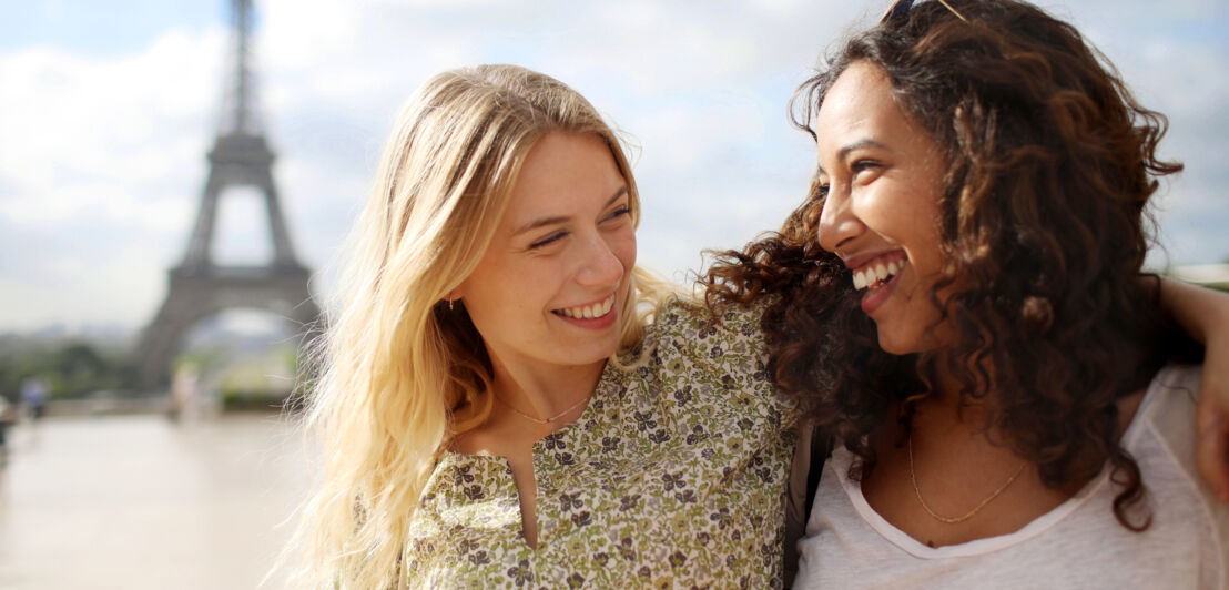 Porträt von zwei jungen Frauen, die lachend Arme umeinander legen, im Hintergrund unscharf die Silhouette des Eiffelturms.