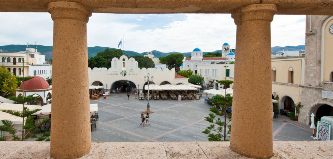 Blick durch die Säulen einer Empore hinab auf einen zentralen Platz mit Markthalle und Moschee