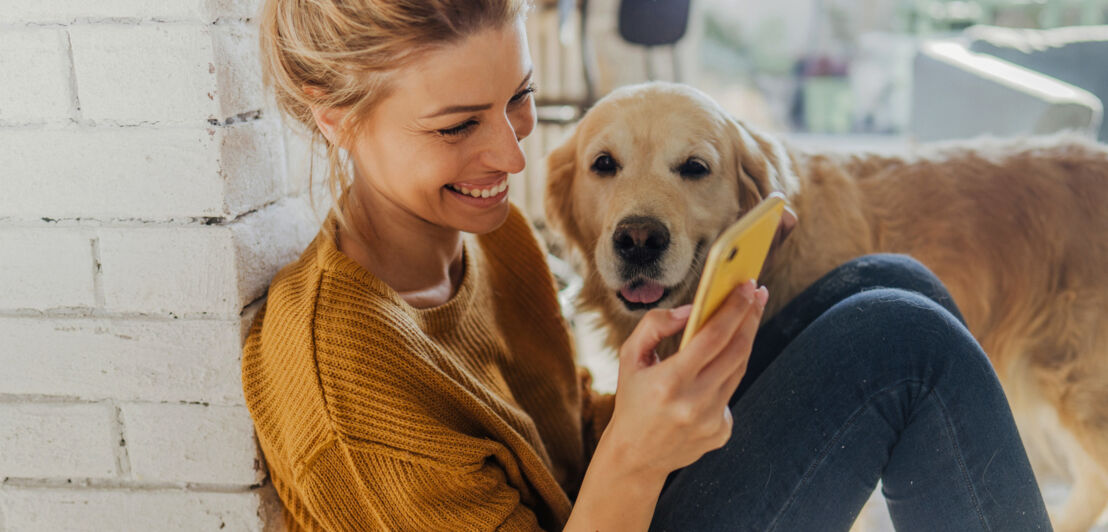 Eine junge Frau sitzt in einer Wohnung neben einem Golden Retriever auf dem Boden und schaut gut gelaunt auf ihr Smartphone.