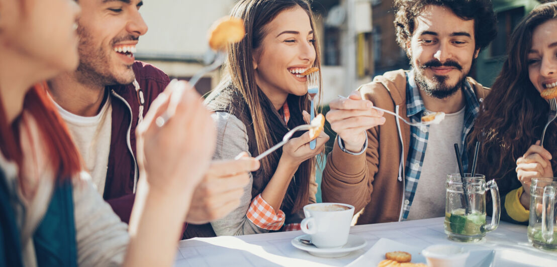 Fünf junge Menschen mit Getränken und Snacks an einem Tisch im Freien.