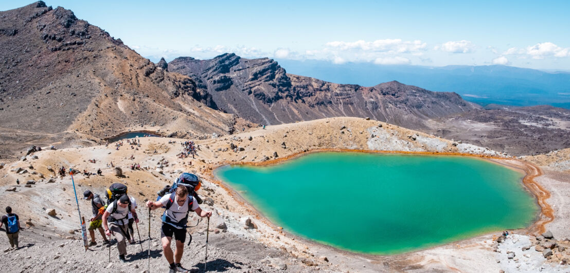 Mehrere wandernde Personen auf einem kargen Berg neben einem grün schimmernden Bergsee