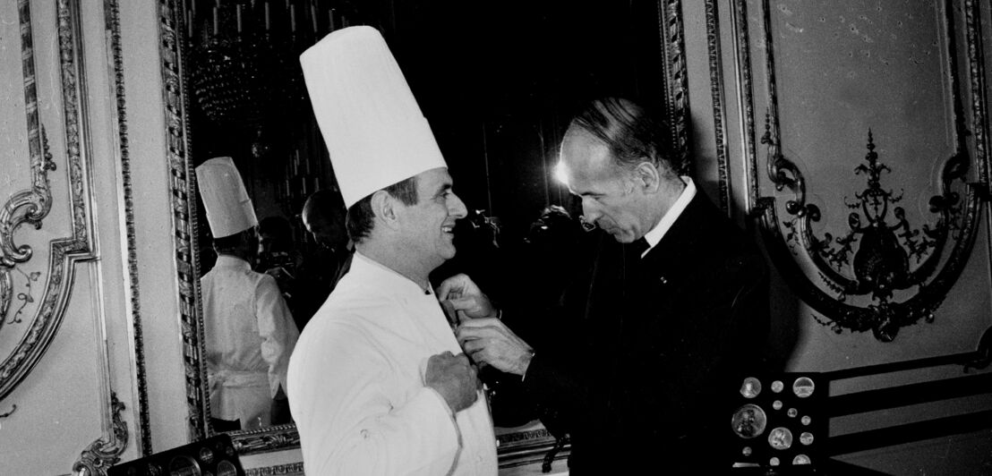 Koch Paul Bocuse bekommt im Beisein von Journalisten einen Orden von Valéry Giscard d’Estaing angesteckt, Schwarzweißaufnahme