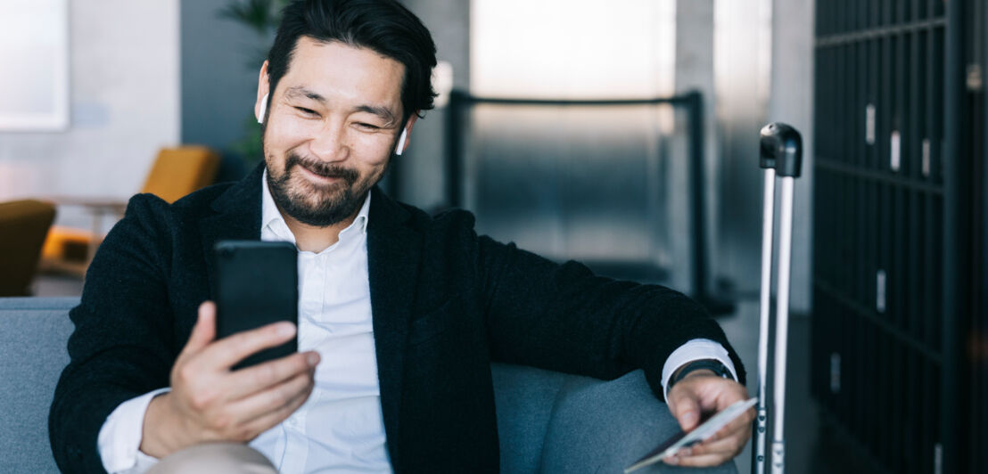 Ein Geschäftsmann mit Reiseunterlagen in einer Hand sitzt auf einem Sofa in einer Lounge und schaut gut gelaunt auf sein Smartphone, neben ihm ein Reisekoffer.