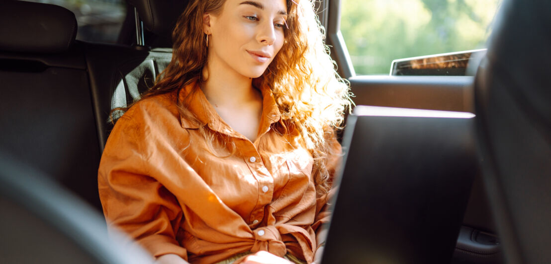 Eine junge Frau arbeitet an ihrem Laptop auf dem Rücksitz eines Autos.