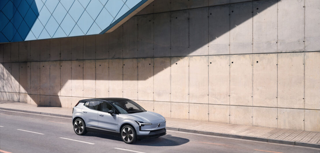 Ein silberfarbener Volvo EX30 fährt auf einer Straße an einer Betonwand vorbei.