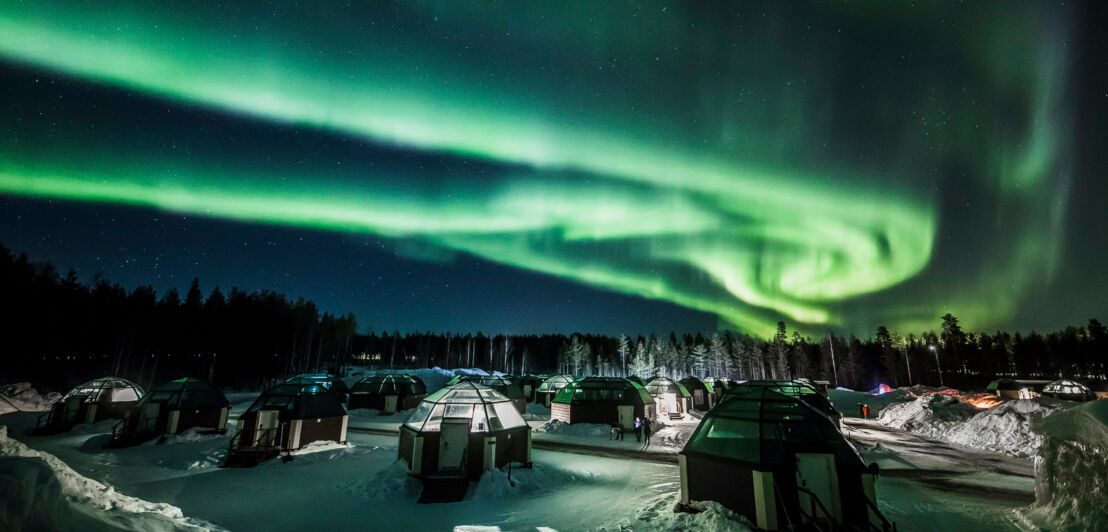 Ein Gelände mit Glasiglus bei Nacht, am Himmel sieht man grüne Polarlichter.