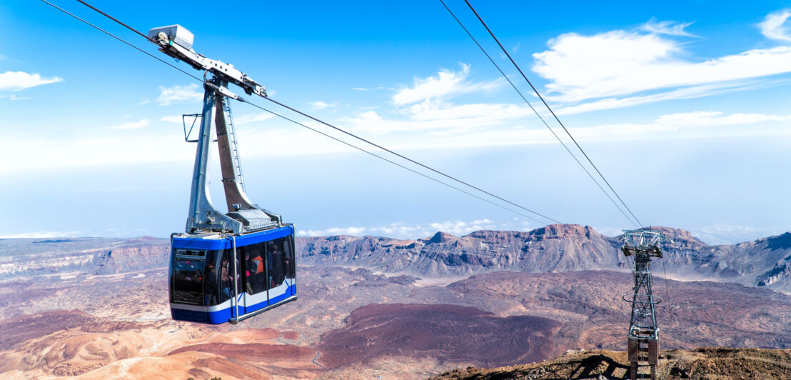 Eine Seilbahn vor karger hügeliger Landschaft, im Hintergrund das Meer.