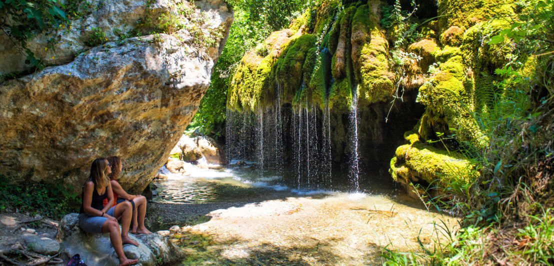 Zwei Frauen sitzen auf einem Stein an einem Wasserfall, umgeben von grüner Natur.