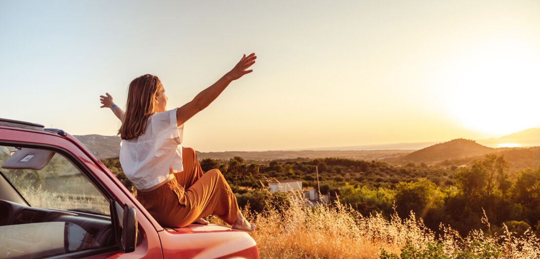 Rückansicht einer jungen Frau mit ausgebreiteten Armen auf der Kühlerhabe eines roten Autos vor einem Landschaftspanorama bei Sonnenuntergang.