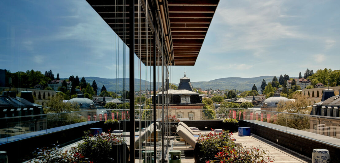 Große Balkonterrasse mit Liegestühlen vor grüner Hügellandschaft, die sich in der Glasfassade des Gebäudes spiegelt.