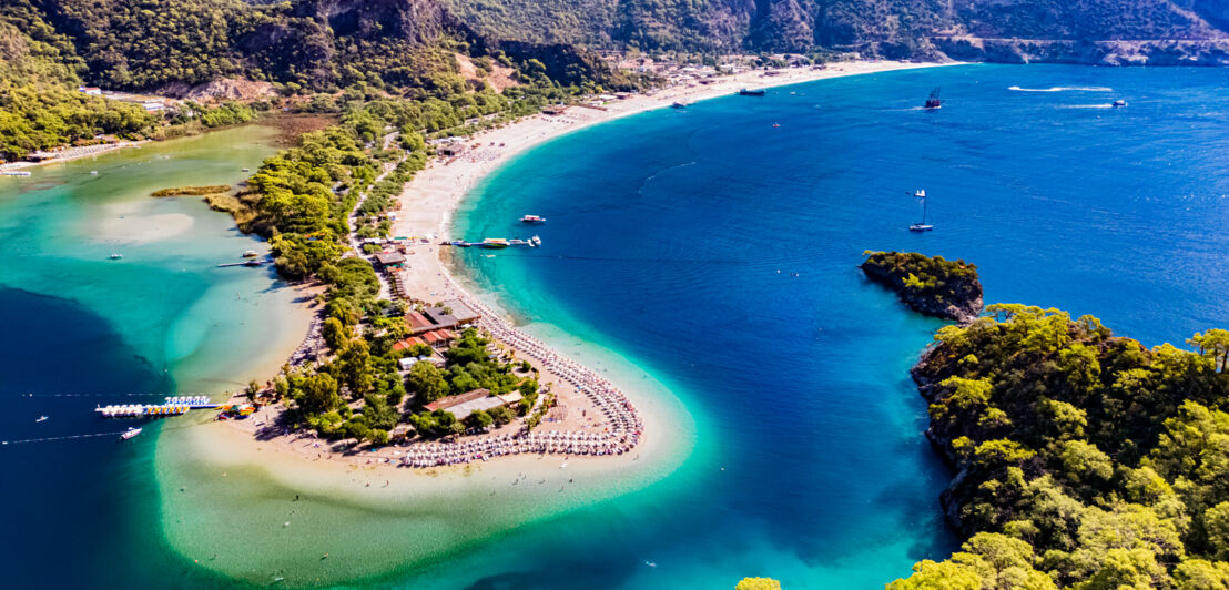 Luftaufnahme einer Landzunge mit weißem Sandstrand und üppiger Begrünung in einer hügeligen Bucht mit azurblauem Wasser.