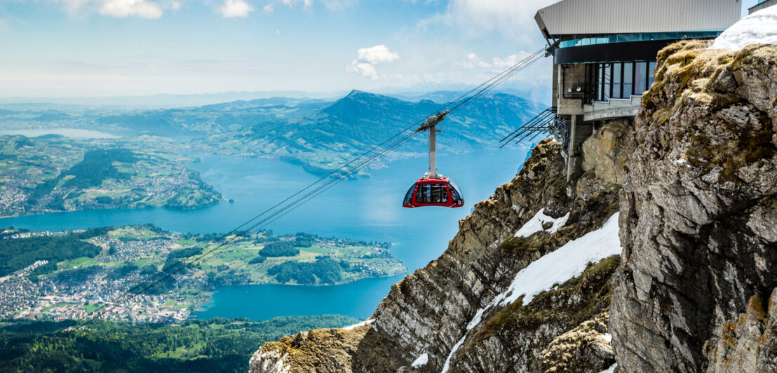 Eine rote Gondel einer Seilbahn an einem Berggipfel vor fjordartiger Landschaft des Vierwaldstättersees aus der Luft.