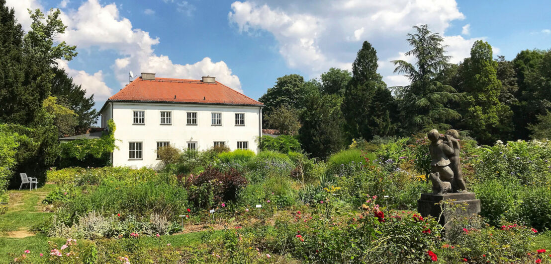 Blühende Rosenbüsche rund um eine kleine Statue in einem Rosengarten, im Hintergrund eine weiße Villa.