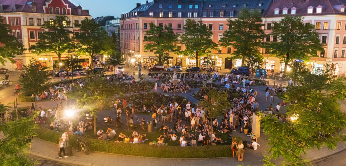 Ein belebter, runder Stadtteilplatz, umgeben von Altbauten am Abend.