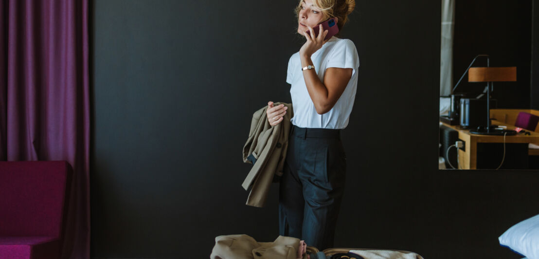 Eine Frau in einem Hotelzimmer, die ihr Smartphone ans Ohr hält und in der anderen Hand eine Jacke hält. Auf dem Bett liegt ein geöffneter Koffer mit verschiedenen Klamotten. 