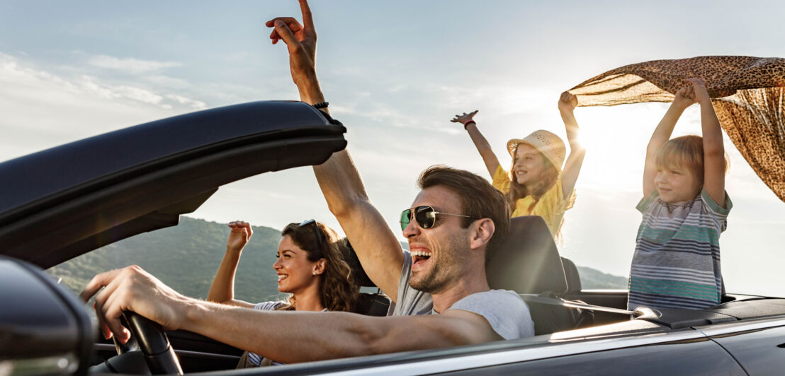 Eine Familie mit zwei Kindern fährt mit ausgestreckten Händen in einem Cabriolet bei Sonnenschein.