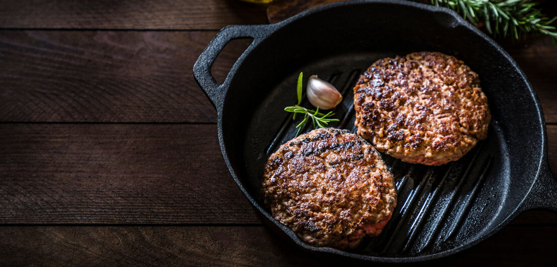 Kochpfanne mit zwei gegrilltem Burgerpattys, umgeben von Rosmarin und einer Olivenölflasche auf einem Holztisch.