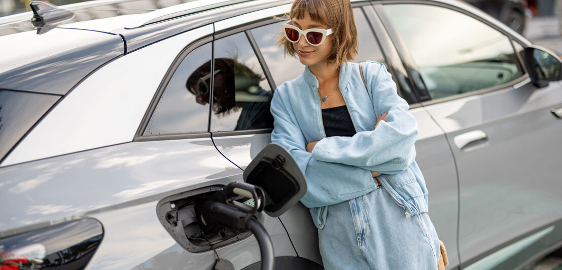 Portrait of a young woman standing near her electric car charging on a public station. Concept of modern lifestyle and success