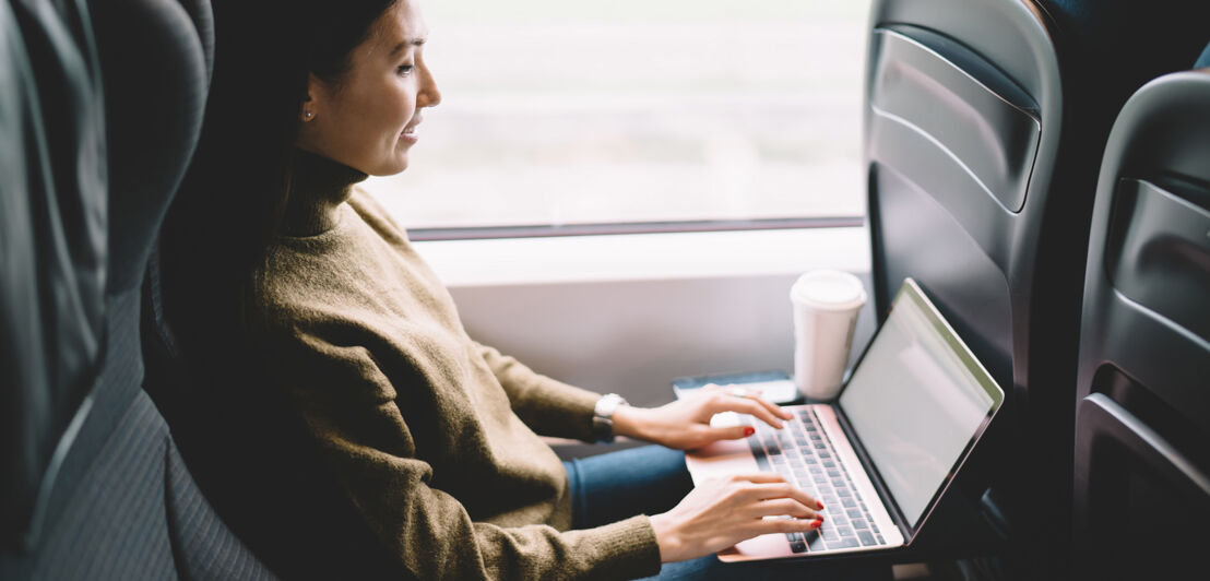 Side view of optimistic female freelancer in casual wear browsing laptop while resting with coffee in train on gray day