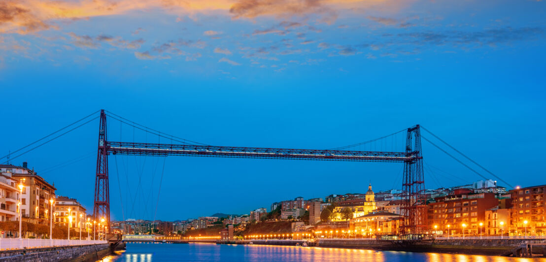 Die Puente de Vizcaya ragt bei Nacht über den Fluss, rechts und links befinden sich beleuchtete Häuser.