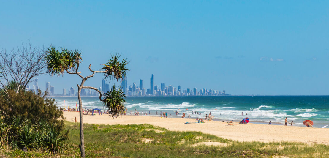 Belebter, weitläufiger Sandstrand am welligen Meer, im Hintergrund eine Skyline.