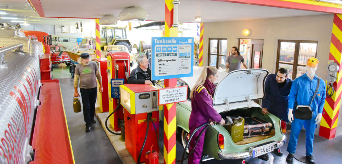 Personen an einer nachgebildeten Tankstelle mit Oldtimern in einem Ausstellungsraum.