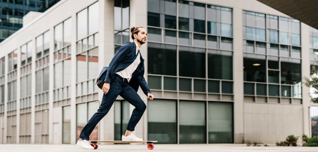 Ein junger Mann im Anzug mit weißen Sneakern auf einem Skateboard vor einem Bürogebäude.