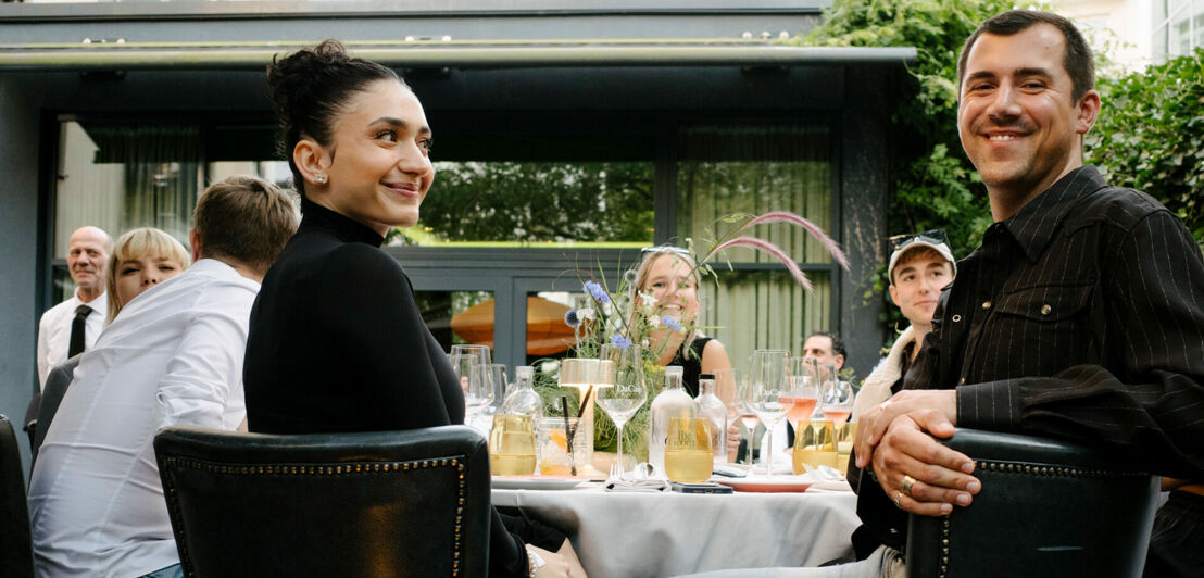 Mehrere Personen sitzen an einem Esstisch auf einer Hotelterrasse, zwei Personen im Vordergrundsind sind lächelnd in Richtung Kamera gewandt.