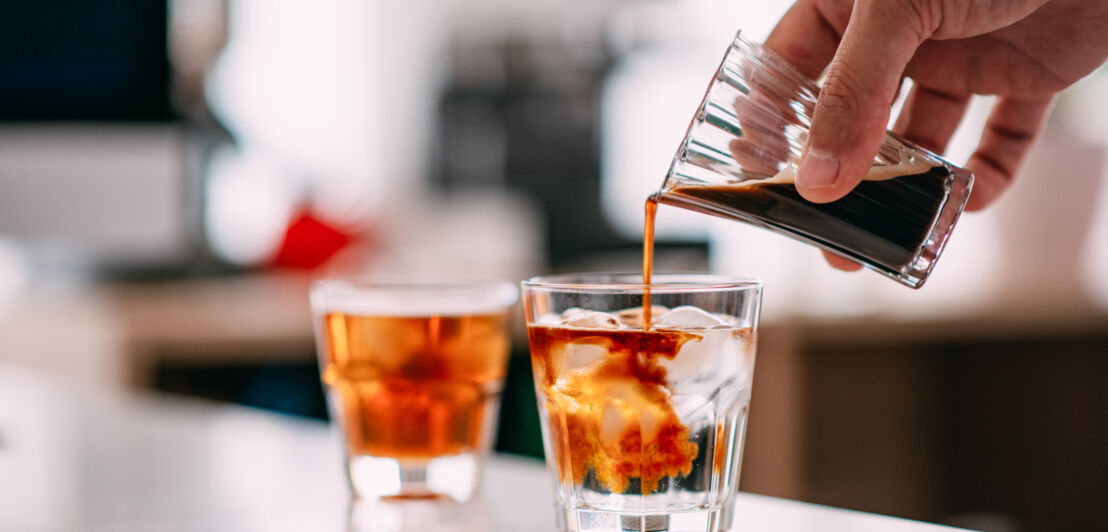 Eine Hand, die Espresso aus einem Glas in ein anderes Glas mit Wasser und Eiswürfeln schüttet.