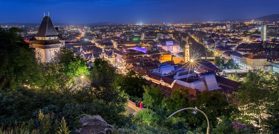 Panoramablick auf Graz von einem begrünten Berg mit Uhrenturm bei Nacht.