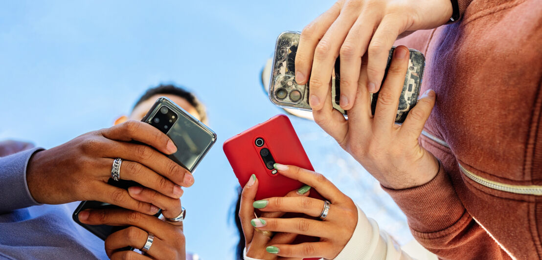 Untersicht dreier Smartphones in den Händen von drei Personen vor blauem Himmel.