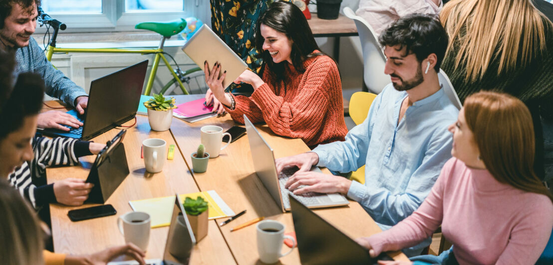 Eine Gruppe junger Leute sitzt zusammen an einem Tisch im Coworking Space und arbeitet mit Laptops