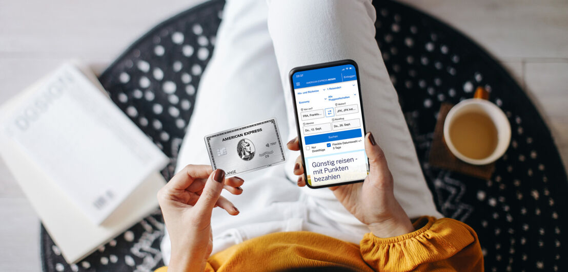 Aufsicht einer sitzenden Person mit silberner Kreditkarte von American Express und Smartphone mit Amex Flugbuchungsmaske auf dem Display in ihren Händen.